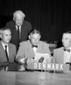 Three men sitting beside eachother at a table with the sign "DENMARK". Behind them stands an elderly man