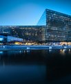 Taken at night. Beautiful evening view of an asymmetric building placed just by the water. In the windows, there are lights.