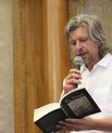 A waist-up picture of Karl Ove Knausgård. He is wearing a white shirt and has grey hair and a grey beard. In one hand he is holding a book with the title "My Struggle" and in the other hand he has a microphone lifted to his mouth.