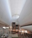 Inside of a church/a chapel, very white coloured. Looks quite closed with not much space since the walls on the sides are arching in.At the back theres an organ. There are also rows of benches to sit.