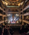 Inside of a theatre, large round shaped seatings with a large crowd looking at the scene. The hall is filled with lights and colours.