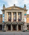 A large bulding in a light beige colour. Has the title 'NATIONALTHEATER' engraved into the building. There is a sculpture of two men standing to the right and left of the building.