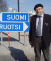 Elder man standing beside two road signs. The first sign says Suomi and the other sign says Ruotsi