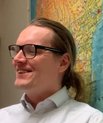 A smiling young man with long hair in a ponytail wearing glasses sitting in front of a world map