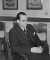 Black and white portrait of a man with glasses sitting in a chair in a suit